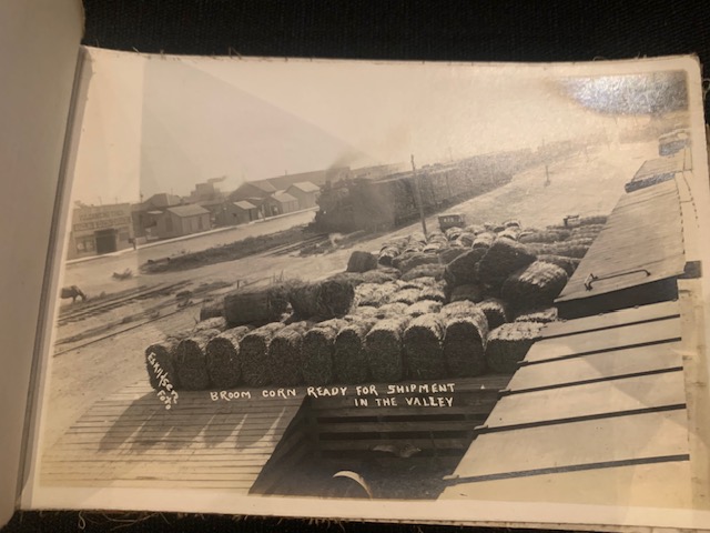 Bales of broom corn on a loading dock, with a steam train waiting on the tracks in the background.